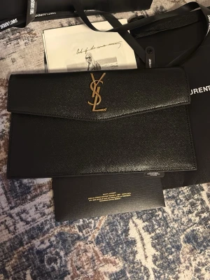 YSL väska - Säljer en svart kuvertväska från Saint Laurent med klassisk YSL-logga i guld på framsidan. Väskan är tillverkad i skinn och har en stilren, rektangulär form med magnetstängning och innerfack. Perfekt för att lyfta din outfit.