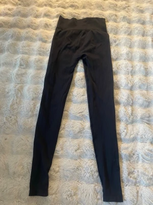 Svarta ribbade leggings med hög midja - Säljer ett par svarta leggings med ribbad struktur och hög midja. De är stretchiga och sitter tajt längs benen, perfekta för en trendig och bekväm look. Materialet känns mjukt och följsamt. Märke och storlek är okänt.