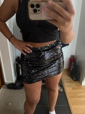 Svart paljettkjol i kort modell - Snygg svart kortkjol täckt med glittriga paljetter. Kjolen har en tight passform och elastisk midja, perfekt för att sticka ut. Passar dig som vill ha en trendig och festlig look.