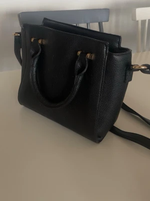 Svart handväska i läder med axelrem - Stilren svart handväska i strukturerat läder med både handtag och justerbar axelrem. Väskan har guldfärgade metalldetaljer och en boxig form som ger den en modern look. 
