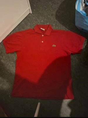 Röd pikétröja från Lacoste - Klassisk röd pikétröja från Lacoste med den ikoniska krokodilloggan på bröstet. Tröjan har korta ärmar, krage och knäppning med två knappar framtill. Perfekt för en stilren och sportig look.
