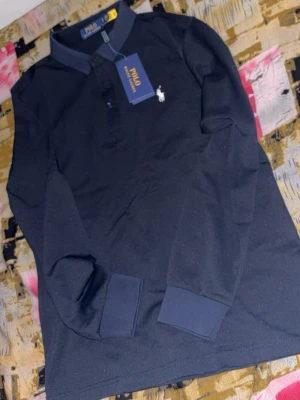 Mörkblå långärmad Polo Ralph Lauren - Klassisk mörkblå långärmad skjorta från Polo Ralph Lauren med vit broderad logga på bröstet. Skjortan har krage, knappar framtill och ribbade muddar vid ärmsluten. Tillverkad i mjuk bomull för en bekväm känsla.