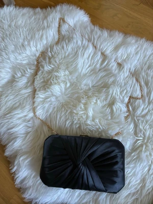 Gina Tricot Svart kuvertväska med guldkedja - Snygg svart kuvertväska i satin med elegant draperad front och guldfärgad kedja som axelrem. Väskan har en rektangulär form och stängs med ett metallspänne upptill. Perfekt accessoar för att lyfta din outfit.