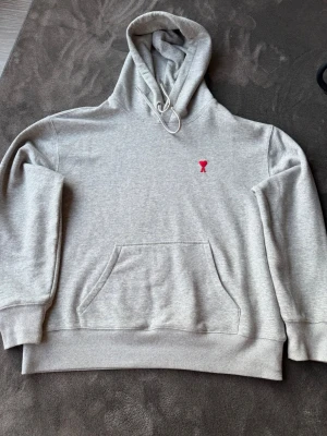 Grå hoodie från Ami Paris med röd logga - Snygg grå hoodie från Ami Paris med den ikoniska röda hjärt-loggan broderad på bröstet. Hoodien har huva med dragsko och en stor magficka framtill. Tillverkad i mjuk bomullsblandning och har ribbade muddar vid ärmslut och nederkant.