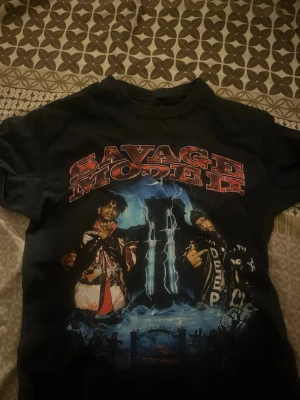 Svart Savage Mode II t-shirt - Cool svart t-shirt med stort Savage Mode II-tryck framtill och färgstark grafik. T-shirten har korta ärmar och en klassisk rund halsringning. Perfekt för dig som gillar streetwear och statement-plagg.