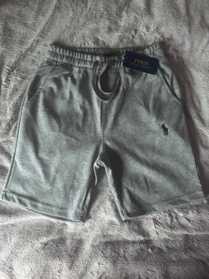 Grå shorts från Polo Ralph Lauren - Snygga grå shorts från Polo Ralph Lauren med elastisk midja och snörning. Klassisk logga broderad på ena benet. Perfekta för sommaren och tillverkade i mjuk bomull för maximal komfort. Dom är i stl M men små i storleken, kan passa S och xs beroende på hur du vill ha passformen. Pris kan diskuteras!