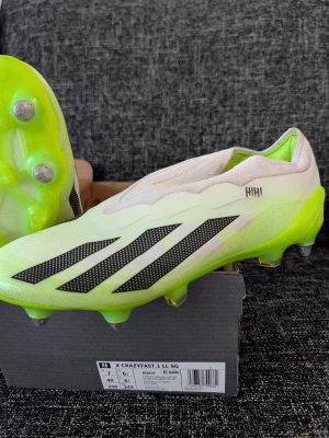 Adidas X Crazyfast.1 LL SG fotbollsskor - Säljer ett par Adidas X Crazyfast.1 LL SG fotbollsskor i vitt och neongrönt med svarta detaljer. Skorna har låg vikt, strumpliknande passform och metall-dobbar för spel på mjuka gräsplaner. Snygg design med tre svarta ränder på sidan och modern look.