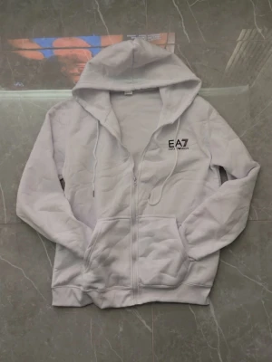 Vit hoodie från EA7 Emporio Armani - Snygg vit hoodie med dragkedja från EA7 Emporio Armani. Hoodien har justerbar huva med snören, två stora fickor framtill och en stilren logga på bröstet. Perfekt för en chill och sportig look.
