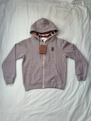 Grå zip-hoodie från Burberry - Snygg grå hoodie med dragkedja från Burberry. Klassisk huva med ikoniskt rutigt foder och dragsko. Bröstet pryds av Burberrys TB-logga i svart. Mjuk bomullsblandning och två fickor framtill. Perfekt för en stilren och avslappnad look.