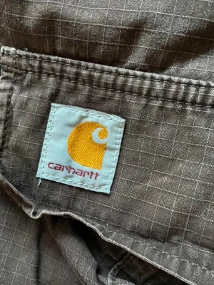 Grå cargobyxor från Carhartt - Säljer ett par grå cargobyxor från Carhartt med rutigt mönster i tyget och klassisk knapp med logga. Byxorna har flera fickor och är tillverkade i slitstarkt material, perfekta för en avslappnad streetstil.