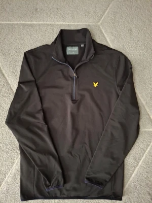 Svart Lyle & Scott tröja med dragkedja - Svart långärmad tröja från Lyle & Scott med halv dragkedja och klassisk gul logga på bröstet. Tröjan har hög krage och är tillverkad i ett mjukt, stretchigt material som känns bekvämt mot huden. Perfekt för en stilren och sportig look.