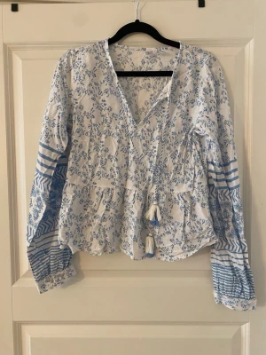 Blommig blus från Rainbow med tofsar - Söt blus från Rainbow i vitt med blått blommönster och randiga detaljer på ärmarna. Långärmad modell med v-ringning och dekorativa tofsar framtill. Luftigt material, perfekt för en avslappnad och trendig look.
