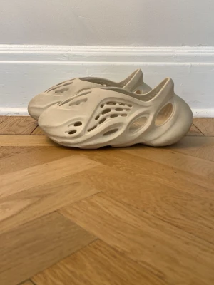 Beige Yeezy Foam Runner tofflor - Säljer ett par beige Yeezy Foam Runner tofflor med futuristisk design och många hål för ventilation. Tillverkade i lätt och bekväm EVA-skum som formar sig efter foten. Perfekta för chill dagar hemma eller när du vill sticka ut med din stil.