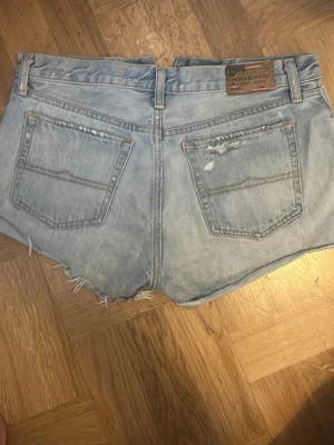 Ljusblå jeansshorts från Ralph Lauren - Säljer ett par ljusblå jeansshorts från Denim & Supply Ralph Lauren. Shortsen har råa kanter, slitna detaljer och klassiska bakfickor. Perfekta för sommaren och har en avslappnad vibe.