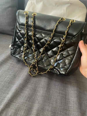 Svart quiltad axelväska med kedja - Snygg svart axelväska i glansigt quiltat material med guldfärgade kedjor som axelrem. Väskan har ett lock och röd insida med dragkedjefack. Perfekt för dig som gillar klassisk men trendig stil.
