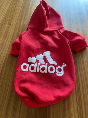 Röd hundtröja med huva Adidog L: 35cm - Röd hundtröja med huva och tryckknappar framtill. Tröjan har ett vitt tryck med texten 'adidog' och ett benmotiv på ryggen. Tillverkad i mjukt tyg, perfekt för att hålla din hund varm och snygg under promenader. Ryggläng: 35cm.