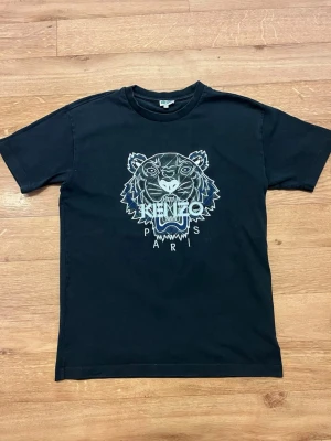 Svart Kenzo t-shirt med tigertryck - Svart t-shirt från Kenzo med ikoniskt tigertryck och Kenzo Paris-logga på bröstet. Klassisk passform med rund hals och korta ärmar. Perfekt för dig som vill sticka ut med en stilren och trendig look.