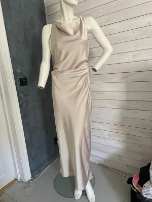 Champagnefärgad långklänning i satin - Elegant långklänning i champagnefärgad satin med draperad halsringning och ärmlös design. Klänningen har en smickrande passform och ett mjukt, glansigt material som ger en lyxig känsla. Perfekt för dig som vill sticka ut med en stilren look.