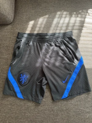 Svarta Nike shorts med blå detaljer - Säljer ett par svarta Nike shorts i storlek Medium med blå detaljer och Nederländernas landslagsmärke på vänster ben. Shortsen har elastisk midja, två fickor med dragkedja och är tillverkade i ett lätt och sportigt material. Perfekta för dig som gillar sportig stil.