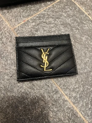 Svart kortfodral från Saint Laurent - Snyggt svart kortfodral från Saint Laurent i präglat läder med guldfärgad YSL-logga framtill. Fodralet har flera kortfack och kommer med originalask. Perfekt accessoar för dig som vill ha något stilrent och lyxigt i fickan.
