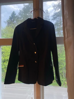 Svart ridkavaj med rödvit detalj - Snygg svart ridkavaj med klassisk krage och tre knappar framtill. På ena fickan finns en stilren detalj med röd, vit och blå rand. Perfekt för tävling eller träning inom ridsport. Långärmad och figursydd modell. Aldrig använd
