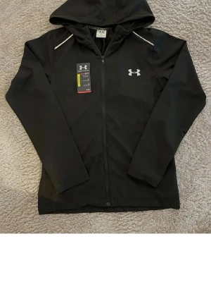 Svart hoodie från Under Armour - Svart hoodie från Under Armour med dragkedja och huva. Framtill finns en vit logga på bröstet och baktill står det 'UNDER ARMOUR' i vitt. Tillverkad i ett sportigt, lätt material med långa ärmar och diskreta vita detaljer vid axlarna.Säljer på grund att den är litten på mig.