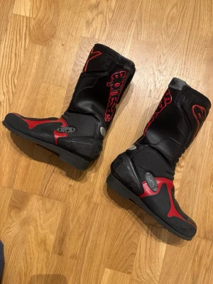 Svarta och röda MC-stövlar Alpinestars - Säljer ett par svarta och röda motorcykelstövlar från Alpinestars i storlek 36-37 som har lite slitage men fungerar bra och är hela. Stövlarna har högt skaft, robust sula och detaljer i rött med logga på sidan och framtill. Tillverkade i slitstarkt syntetmaterial och läder, perfekta för dig som gillar att köra hoj.