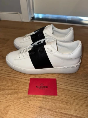 Valentino Garavani vit/svart skor - Stilrena vita sneakers från Valentino Garavani med en bred svart läderdetalj över mitten. Klassisk låg modell med vita snören och diskreta nitar på hälen. Perfekta för dig som gillar lyxiga och trendiga skor med minimalistisk design.