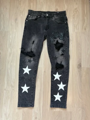 Svarta Amiri jeans med stjärnor och slitningar - Unika svarta jeans med stora vita stjärnor på benen, slitna detaljer och hål på låren samt broderade bokstäver i svart och grått. Jeansen har en smal passform och klassisk femficksdesign. Perfekta för dig som gillar streetstyle och att sticka ut ,amiri-jeans, rymliga framfickor AMR-textilier.