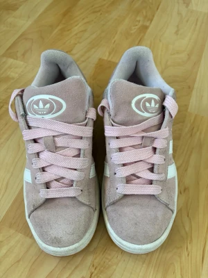 Adidas campus rosa sneakers i mocka  - Säljer ett par snygga Adidas sneakers i ljusrosa mocka med vita detaljer och klassiska tre ränder på sidan. Skorna har rosa skosnören och Adidas-logga på plösen. Låg modell som passar perfekt till streetwear och vardagsstil.