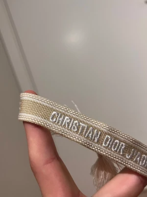 Beige broderat armband Dior - Snyggt beige armband från Christian Dior med texten 'CHRISTIAN DIOR J'ADIOR' broderad i vitt. Armbandet är gjort i textil och har en stilren, bohemisk känsla med tofsar i ändarna. Perfekt accessoar för att lyfta din outfit.