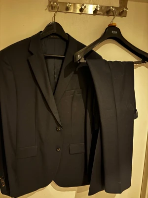 Svart kostym från BOSS - Snygg svart kostym från BOSS med klassisk kavaj och matchande byxor. Kavajen har två knappar, fickor med lock och en bröstficka. Perfekt för dig som vill ha en stilren och tidlös look. Klockren nu inför val/student!     OBS!!!!   Storlek kavaj: 42E    Storlek kostymbyxor: 40L