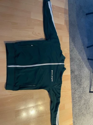 Grön zip-hoodie från Palm Angels - Mörkgrön zip-hoodie från Palm Angels med vita detaljer längs dragkedjan och på ärmarna. Tröjan har två fickor med dragkedja framtill och tryckt logga på bröstet. Perfekt för en sportig och avslappnad stil.