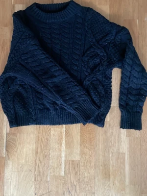 Svart kabelstickad tröja - Mysig svart kabelstickad tröja med rund halsringning och grovstickat mönster. Tröjan har långa ärmar och ribbade muddar vid ärmslut och nederkant. Perfekt för lager-på-lager och en cool vardagslook.
