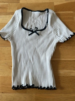 Ribbad topp med rosett och svarta detaljer - Supersöt ribbad topp i ljus färg med svarta kanter och liten rosett vid den fyrkantiga urringningen. Kortärmad och figurnära passform, perfekt för en trendig look.