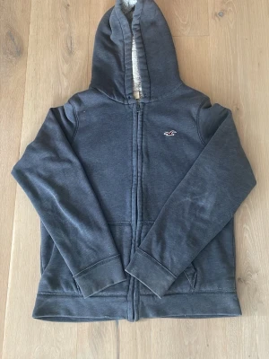 Mörkgrå hoodie från Hollister - Mysig mörkgrå hoodie från Hollister med dragkedja och vit fuskpälsfoder på insidan. Klassisk logga på bröstet och två fickor framtill. Huva med mjukt foder för extra komfort.💕