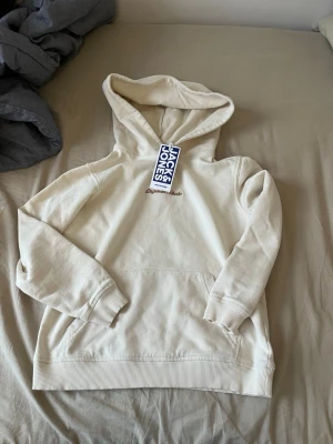 Vit hoodie från Jack & Jones - Snygg vit hoodie från Jack & Jones med broderad logga på bröstet. Klassisk modell med huva och känguruficka framtill. Mjuk bomullskänsla och ribbade muddar vid ärmslut och nederkant. Perfekt för en avslappnad och stilren look.