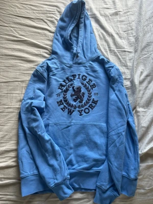 Blå hoodie från Hilfiger New York - Snygg ljusblå hoodie från Hilfiger New York med broderad svart logga och lejon på bröstet. Klassisk känguruficka framtill och huva. Perfekt för en chill och stilren look. Mjuk bomullskänsla och ribbade muddar vid ärmslut och nederkant.