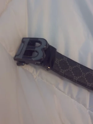 Svart mönstrat bälte med B-spänne - Snyggt svart bälte med ett stort B-format metallspänne och diskret mönster med små B:n längs hela bandet. Perfekt accessoar för att lyfta din outfit med en exklusiv touch. Bältet har en modern och stilren design.PRIS GÅR ATT DISKUTERA 