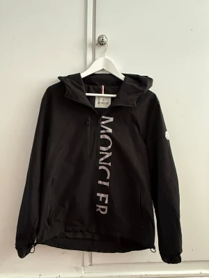Svart vindjacka från Moncler - Svart vindjacka från Moncler med huva och dragkedja. Stor vit Moncler-logga längs dragkedjan framtill och klassisk logotyp på vänster ärm. Jackan har justerbara snören i nederkant och är tillverkad i ett lätt, vindtätt material. Strl:S✅ Pris kan diskuteras!💸 Hör av dig om du har några funderingar!👍