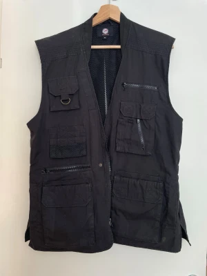 Svart utility-väst med många fickor - Svart utility-väst med flera praktiska fickor, dragkedjor och meshdetaljer. Västen har justerbara band i sidorna och är tillverkad i ett slitstarkt tyg. Lite oversized 