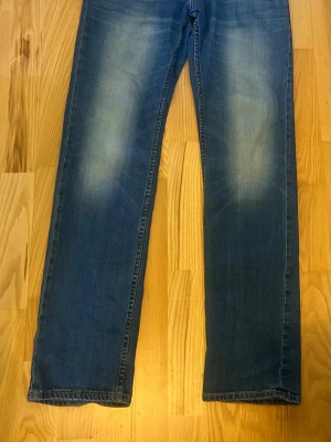 Blå raka jeans från Lee - Snygga blå jeans från Lee i storleken 31/34 med raka ben och klassisk femficksdesign. Jeansen har en lätt tvättad look med slitningar på låren och orangea sömmar. Perfekta för en avslappnad stil och tillverkade i mjukt denimtyg.