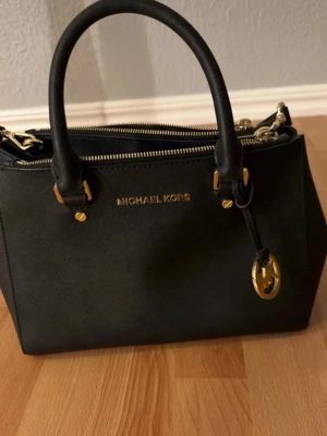 Svart handväska från Michael Kors - Snygg svart handväska från Michael Kors i strukturerat läder med guldfärgade detaljer och dubbla handtag. Väskan har dragkedja upptill och ett dekorativt MK-hänge. Perfekt för dig som gillar stilrena accessoarer.