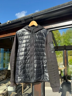 Vindjacka Peak Performance - Svart vindjacka från Peak Performance med quiltad framsida och huva, perfekt för vår eller höst. Jackan har dragkedja framtill, ribbade muddar, logotyp på ärmen och har en sportig look. Ett märke som ni kan se på sista bilden, kanske går att tvätta bort, har ej testat.
