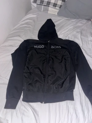 Svart hoodie från Hugo Boss - Snygg svart hoodie från Hugo Boss med dragkedja framtill och broderad logga på bröstet. Hoodien har huva, ribbade muddar och två fickor framtill. Perfekt för en stilren och avslappnad look.