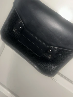 Svart axelväska med kedja - Snygg svart axelväska i konstläder med ormmönstrad struktur på framsidan och detaljer i form av nitar. Väskan har en kedjerem och ett lock med spännande design. Perfekt för dig som vill ha en edgy och trendig look. Pris kan diskuteras