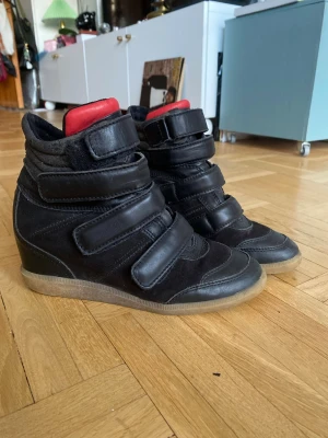 Svarta wedge-pumps med kardborre - Snygga svarta pumps med kilklack och sportig design. Skorna har flera breda kardborreband och detaljer i både mocka och läder. Insidan har en röd vadderad plös som sticker ut. Yttersulan är i gummi. Passar strl 38-39.