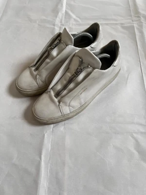 Vita arigato  sneakers med dragkedja - Stilrena vita sneakers från arigato i läder med unik dragkedja framtill istället för klassiska snören. Diskret logga på sidan och svart innersula. Perfekta för dig som gillar minimalistisk och modern design.