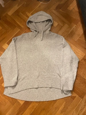 Grå stickad hoodie med snörning - Mysig grå hoodie i stickat material med huva och snörning. Modellen är loose fit med lång ärm och ribbade muddar. 