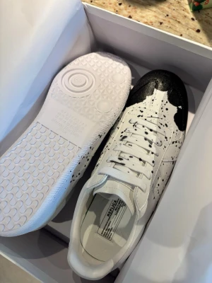 Maison Margiela - Säljer ett par vita sneakers från Maison Margiela med unikt svart färgstänk över tån och sidorna. Skorna har klassisk snörning och är tillverkade i skinn för en lyxig känsla. Perfekta för dig som vill sticka ut med en kreativ och modern look. Kom privat om ni har frågor!